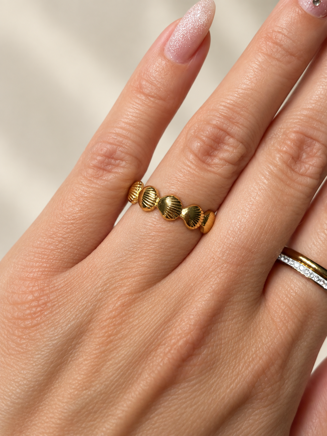 Goldfarbener Ring mit fein gerillten Muschelmotiven am Finger, vor einem weichen cremefarbenen Hintergrund.